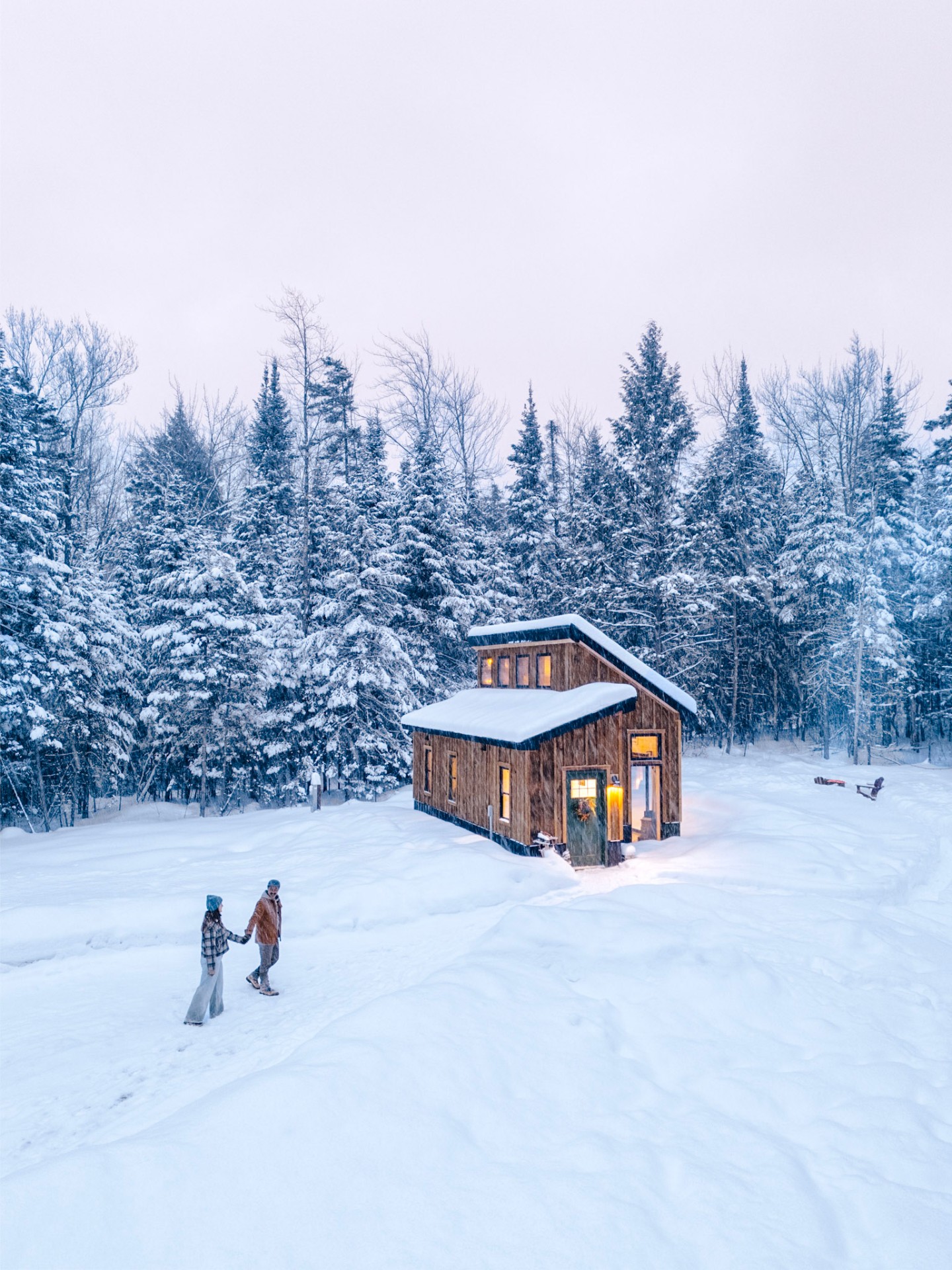 Couple walking hand-in-hand toward a warmly lit wooden tiny house at dusk in a snowy Vermont forest
